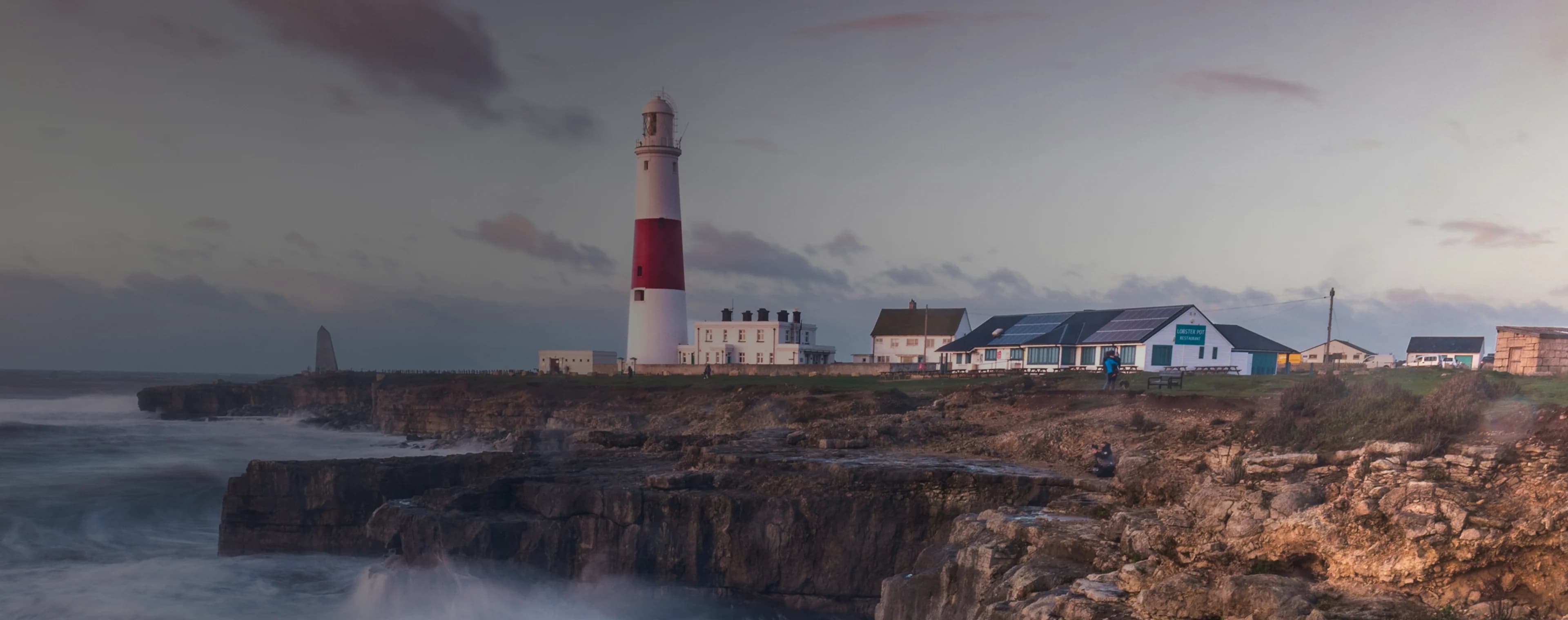 Las olas rompen en un acantilado británico con un faro en la distancia.