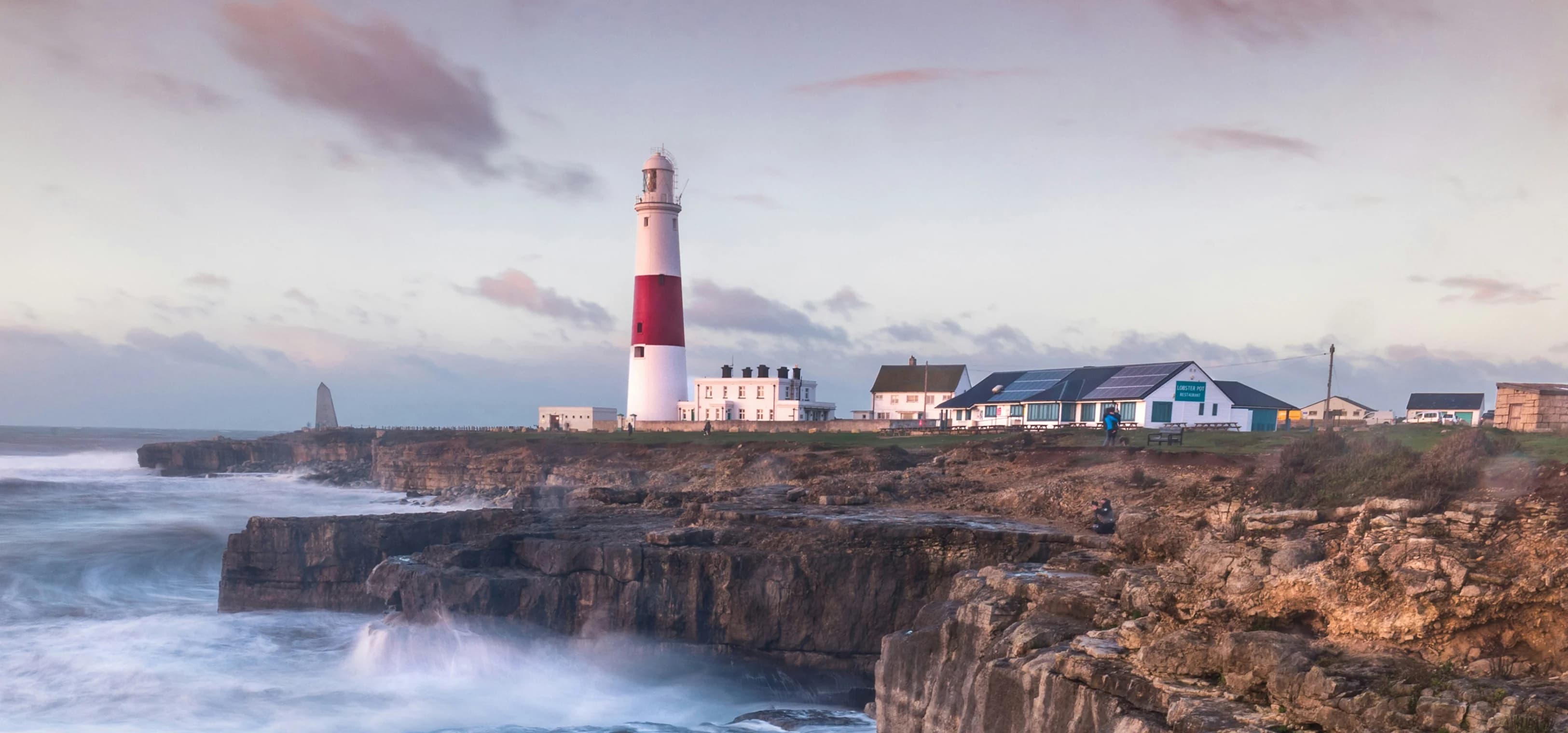 Las olas rompen en un acantilado británico con un faro en la distancia.