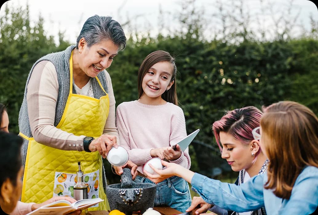 Un adulto enseña a los niños a preparar ingredientes con un mortero durante una actividad práctica de aprendizaje cultural, al aire libre.